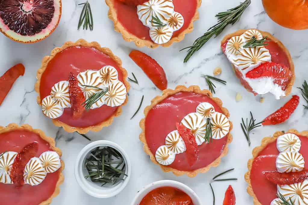 A scene with several rosemary blood orange tarts scattered about with rosemary and blood orange segments lying around