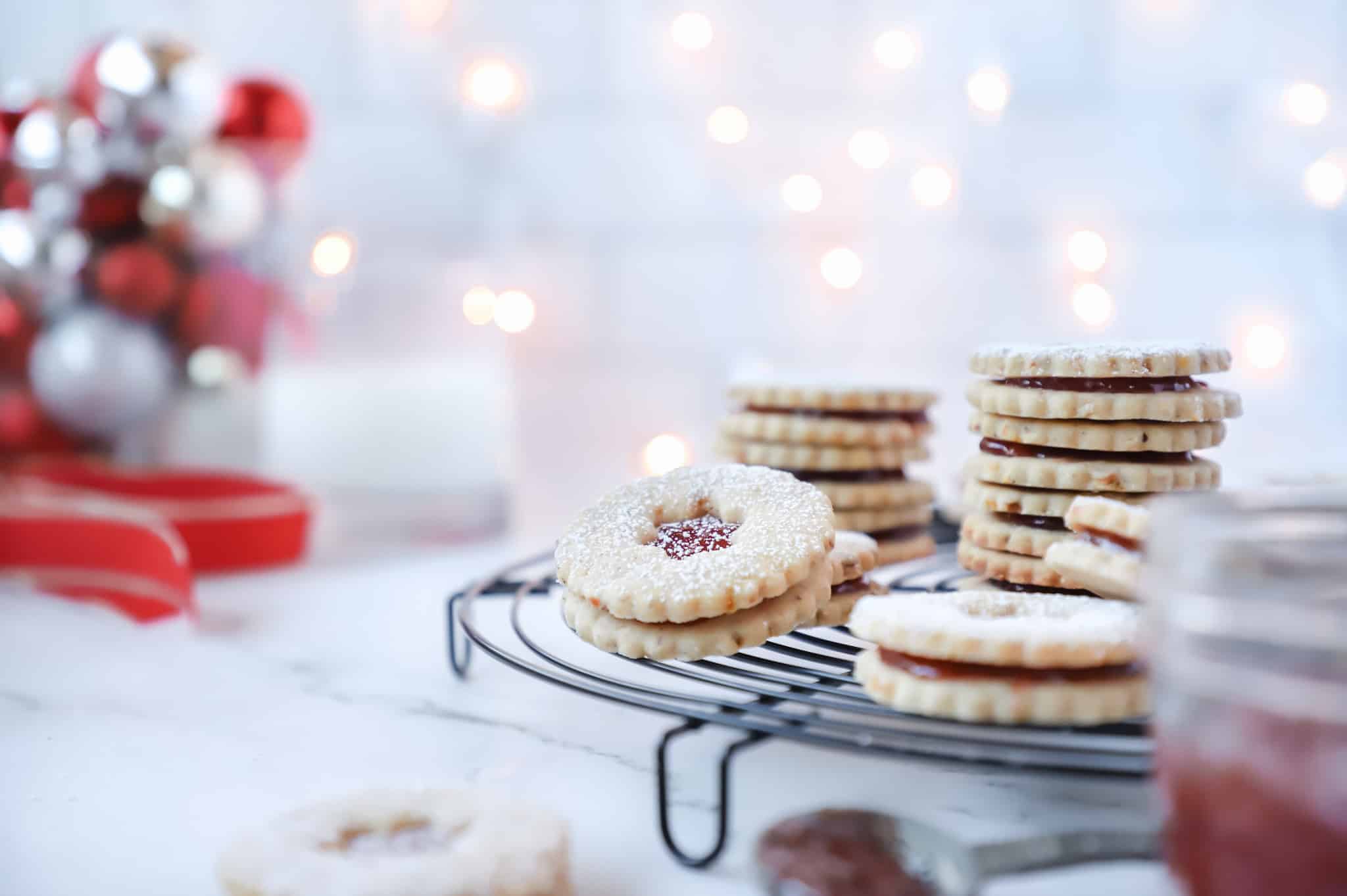 A head on shot of Christmas linzer cookies stacked on a round, black cooling rack with some Christmas lights in the background