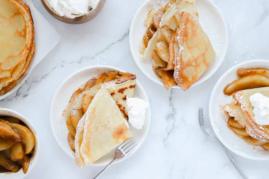 An overhead shot of several plates of caramelized pear crepes with a bowl of whipped cream, extra pears, and extra, unfilled crepes